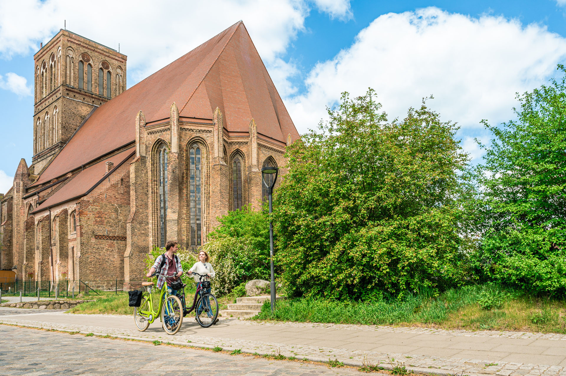 Two cyclists push their bicycles on the sidewalk. Behind them the Nikolaikirche.